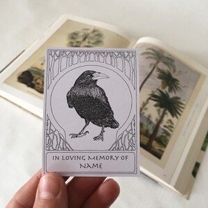 Pu&ograve; includere: Un'illustrazione in bianco e nero di un corvo in una cornice decorativa. Il testo "In loving memory of NAME" &egrave; stampato sotto l'immagine.