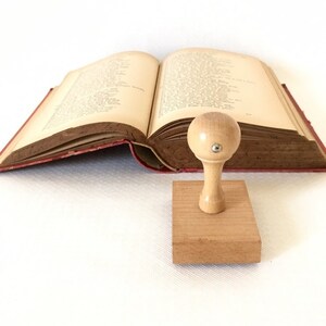 Puede incluir: Un sello de madera con la parte superior redonda y una base rectangular. El sello est&aacute; sobre una superficie blanca delante de un libro abierto.