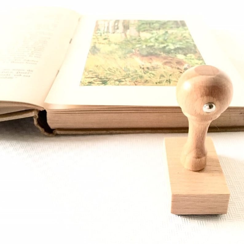 Puede incluir: Un sello de goma de madera con un mango redondo y una base rectangular. El sello est&aacute; sobre una superficie blanca junto a un libro cerrado.