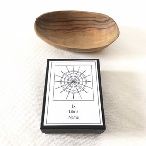 May include: A black box with a white label. The label features a black and white illustration of a compass rose and the text "Ex Libris Name".