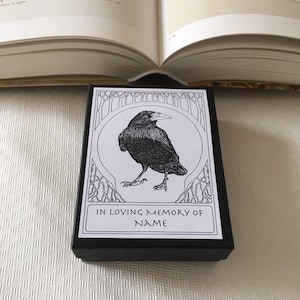 Pu&ograve; includere: Una scatola nera con un'etichetta bianca che presenta un'illustrazione in bianco e nero di un corvo. L'etichetta recita "In memoria affettuosa di NAME".