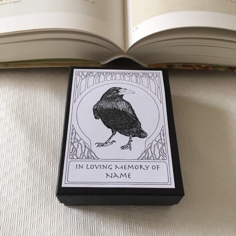 Pu&ograve; includere: Una scatola nera con un'etichetta bianca che presenta un'illustrazione in bianco e nero di un corvo. L'etichetta recita "In memoria affettuosa di NAME".
