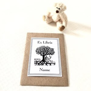 Könnte beinhalten: Ein brauner Papierumschlag mit einem weißen Aufkleber, auf dem "Ex Libris" steht und eine Schwarzweißillustration eines Baumes mit einer darunter sitzenden Person zu sehen ist. Der Text "Name" befindet sich unter der Illustration.