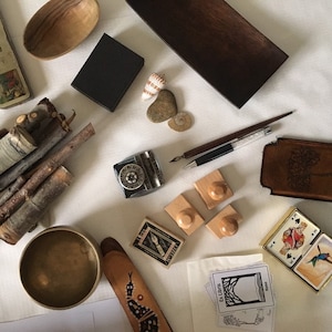 Puede incluir: Una vista cenital de varios objetos, incluyendo un cuenco de madera, un tintero negro, una bandeja de madera, un juego de sellos de madera, una peque&ntilde;a caja de madera, un juego de cartas de juego, una funda de cuero marr&oacute;n, un juego de palos, un cuenco peque&ntilde;o, una cuchara de madera, una concha marina y un bol&iacute;grafo.