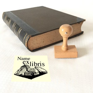 Puede incluir: Un sello de libro de madera con un mango redondo y una base rectangular. El sello presenta una ilustraci&oacute;n en blanco y negro de una pila de libros con el texto "Name ex libris".