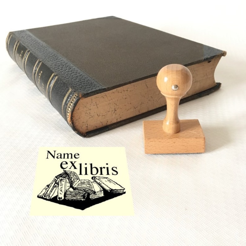 Puede incluir: Un sello de libro de madera con un mango redondo y una base rectangular. El sello presenta una ilustraci&oacute;n en blanco y negro de una pila de libros con el texto "Name ex libris".