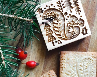 May include: A wooden cookie stamp with a carved squirrel design, surrounded by trees and foliage. The stamp is next to a few stamped cookies and sprigs of evergreen with red berries, all on a wooden surface.