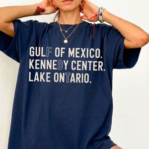 May include: A navy blue short-sleeve t-shirt featuring white text that reads GULF OF MEXICO. KENNEDY CENTER. LAKE ONTARIO. The letters F, D, and T are highlighted in a lighter blue color within the words.