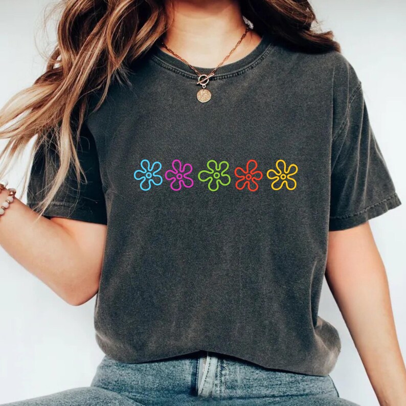 May include: Dark grey t-shirt with five colourful flower designs across the front. The flowers are outlined in blue, purple, green, red, and yellow. The shirt has a washed-out, vintage appearance.