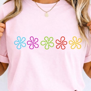 May include: A pale pink t-shirt with five colourful flower designs across the front. The flowers are outlined in blue, purple, green, red, and yellow. The shirt has short sleeves and a round neck.