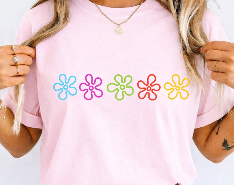 May include: A pale pink t-shirt with five colourful flower designs across the front. The flowers are outlined in blue, purple, green, red, and yellow. The shirt has short sleeves and a round neck.