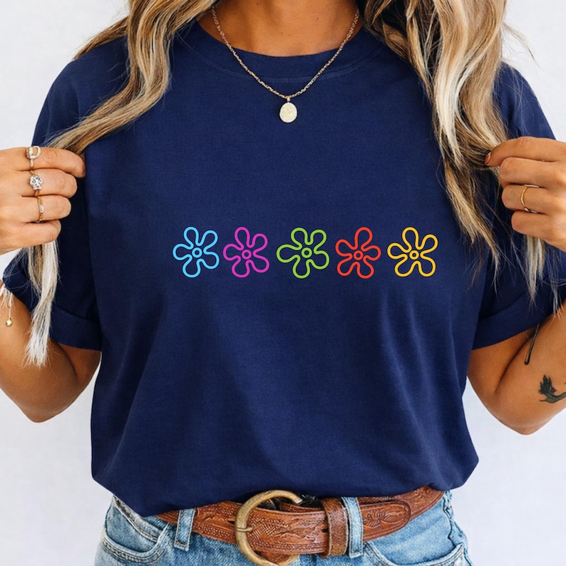 May include: A navy blue t-shirt with a row of five colourful flower designs. The flowers are outlined in black and filled with blue, pink, green, red, and yellow. The shirt is made of a soft material.