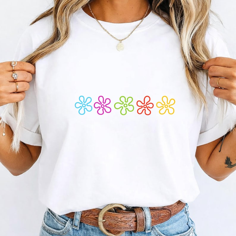 May include: White t-shirt with a row of five colourful flower graphics in blue, pink, green, red, and yellow. The shirt has a crew neck and short sleeves. The design is centred on the front of the shirt.