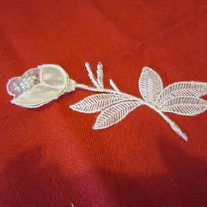 May include: A white lace rose applique with a stem and leaves, embellished with small beads, rests on a red fabric background. The delicate floral design is suitable for embellishing clothing or craft projects.