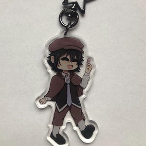 Peut inclure: Un porte-clés en acrylique transparent représentant un personnage de dessin animé aux cheveux bruns, portant un chapeau brun, un manteau noir, une cravate bleue et un pantalon marron. Le personnage tient une sucette rose.