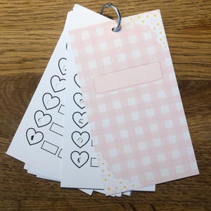 Op de afbeelding: Een stapel papieren kaarten bijeengehouden door een zilveren metalen ring. De kaarten hebben een roze en witte ruitjespatroon, blanco rechthoeken en hartvormen. De kaarten liggen op een houten oppervlak.