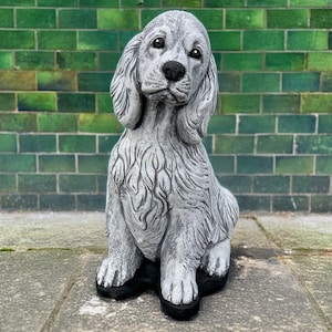 Könnte beinhalten: Graue Betonstatue eines sitzenden Hundes. Der Hund hat lange, hängende Ohren, eine schwarze Nase und eine detaillierte Fellstruktur. Die Statue steht vor einer grünen Backsteinmauer.