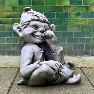 May include: A gray garden gnome statue with a mischievous expression, wearing a pointed hat and tunic. The gnome is seated, with one hand touching its face. The background features a green brick wall.