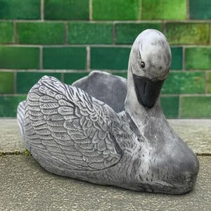Könnte beinhalten: Grauer, schwanförmiger Pflanztopf aus Stein. Der Schwan hat detaillierte Federgravuren an Flügeln und Körper. Der Schnabel ist schwarz, und der Kopf ist nach unten geneigt. Der Pflanztopf steht auf einer Betonoberfläche mit grünem Ziegelhintergrund.