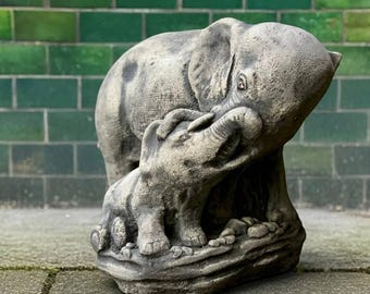 Puede incluir: Una escultura de piedra gris desgastada de un elefante adulto y un elefante bebé. El elefante adulto está posicionado detrás del bebé, con su trompa tocando suavemente la trompa del bebé. La escultura está sobre una base de piedra.