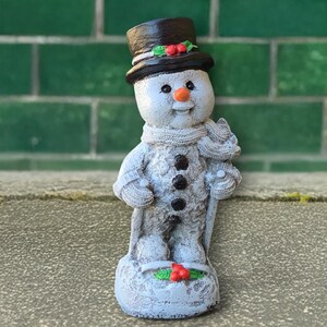 May include: A small, gray snowman figurine with a black top hat, scarf, and coal buttons. The snowman has a carrot nose and is holding a cane. Red berries and green leaves decorate the hat and base. The figurine is standing on a small, snow-covered base.