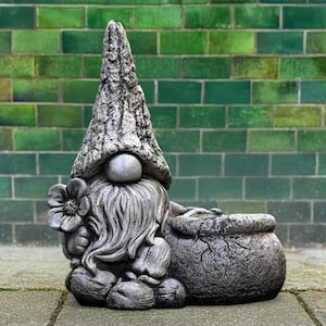 以下が含まれることがあります： 長いひげを生やし、花と袋を持った灰色のコンクリート製庭のノーム像。ノームは背の高いテクスチャード加工の帽子をかぶっています。背景は緑色のレンガの壁です。