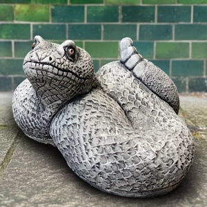 May include: A grey, coiled snake statue with detailed scales and a textured surface. The snake has a detailed head with visible eyes and is positioned on a stone surface, with a green brick wall in the background.