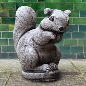 Puede incluir: Estatua de ardilla de hormigón gris en posición sentada, con las patas juntas. La ardilla tiene una cola tupida y una textura de pelaje detallada. La estatua está colocada sobre una superficie de piedra con una pared de ladrillos verdes al fondo.