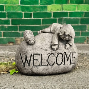 Puede incluir: Una estatua de jardín de piedra gris que representa a un niño descansando sobre una gran roca. La palabra "WELCOME" está tallada en la roca. También hay una figura de caracol en la roca. El fondo es una pared de ladrillos verdes.
