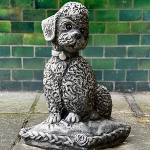 Puede incluir: Una escultura de piedra gris de un caniche sentado en una almohada. El caniche lleva un collar y mira hacia la izquierda. La almohada tiene un ribete de cuerda y un diseño de rosa.