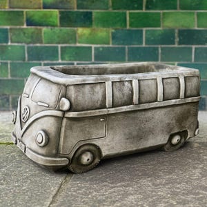 May include: A gray concrete planter shaped like a vintage Volkswagen bus. The planter has a rectangular opening for plants, with details including headlights, wheels, and the VW logo. The background features a green brick wall.