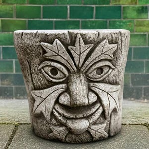 Puede incluir: Una maceta gris, de aspecto envejecido, con un diseño de rostro tallado. La cara presenta detalles de hojas, ojos, nariz y boca sonriente. La maceta está hecha de un material duradero y es adecuada para uso en exteriores. El fondo es una pared de ladrillos verdes.