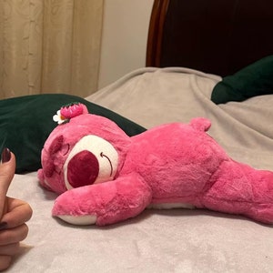 May include: A pink plush bear toy with a white belly and a flower on its head is lying on a bed. The bear has a smiling face and is in a relaxed position. A dark green cushion is behind the bear.