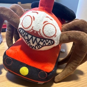 Könnte beinhalten: Ein rotes und schwarzes Plüschspielzeug in Form eines Zuges mit acht braunen Spinnenbeinen. Der Zug hat ein weißes Gesicht mit großen Augen, einem breiten, zahnigen Grinsen und einem schwarzen Hut.