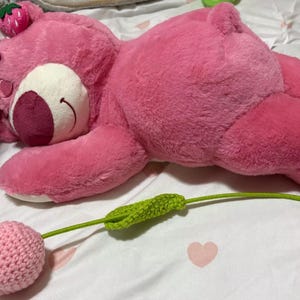 May include: A large pink plush bear with a strawberry on its head, lying on a bed. A crocheted pink flower with a green stem is in the foreground. The bear has white accents and a smiling face.