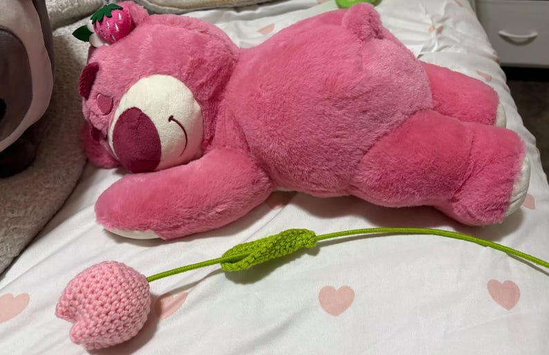 May include: A large pink plush bear with a strawberry on its head, lying on a bed. A crocheted pink flower with a green stem is in the foreground. The bear has white accents and a smiling face.