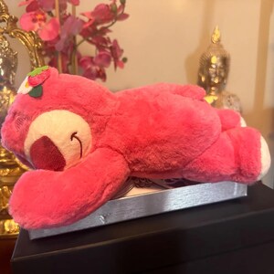 May include: A pink plush bear toy with a strawberry on its head, lying down. The bear has a white belly and a smiling face. The toy is resting on a silver box, with a black surface below.