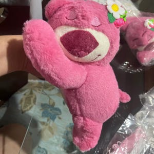 May include: A pink plush bear toy with a white belly and a dark pink nose and mouth. The bear has a flower and strawberry accessory on its head. The bear is a stuffed toy.