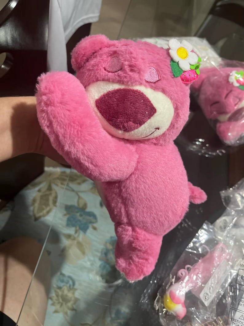 May include: A pink plush bear toy with a white belly and a dark pink nose and mouth. The bear has a flower and strawberry accessory on its head. The bear is a stuffed toy.