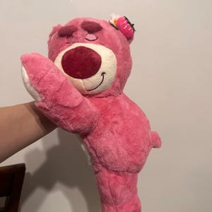May include: A pink plush bear toy with a large red nose and a smiling mouth. The bear has a strawberry and flower accessory on its head. The bear is soft and fluffy, with white accents on its paws.