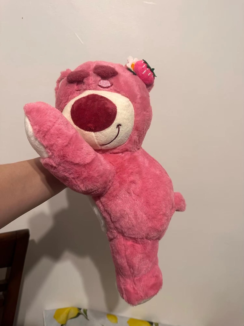 May include: A pink plush bear toy with a large red nose and a smiling mouth. The bear has a strawberry and flower accessory on its head. The bear is soft and fluffy, with white accents on its paws.