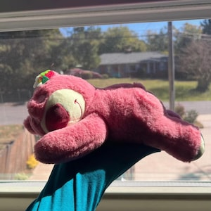 May include: A pink plush bear toy with a red nose and a small green and red floral accent on its head. The bear is lying down, with its eyes closed, and is resting on a blue surface. The toy is in front of a window.