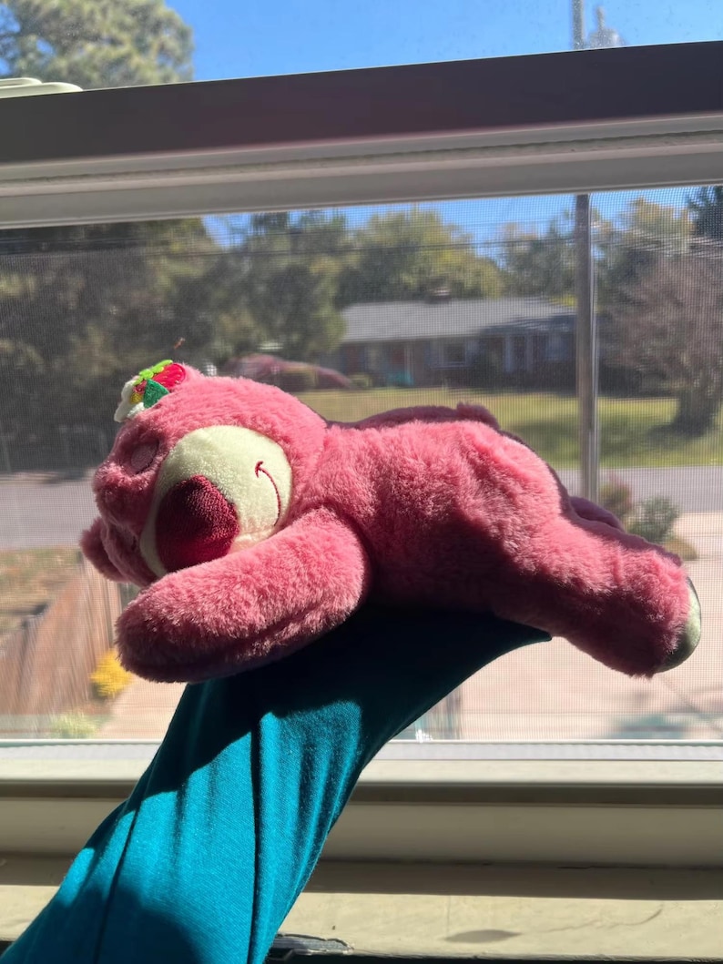 May include: A pink plush bear toy with a red nose and a small green and red floral accent on its head. The bear is lying down, with its eyes closed, and is resting on a blue surface. The toy is in front of a window.