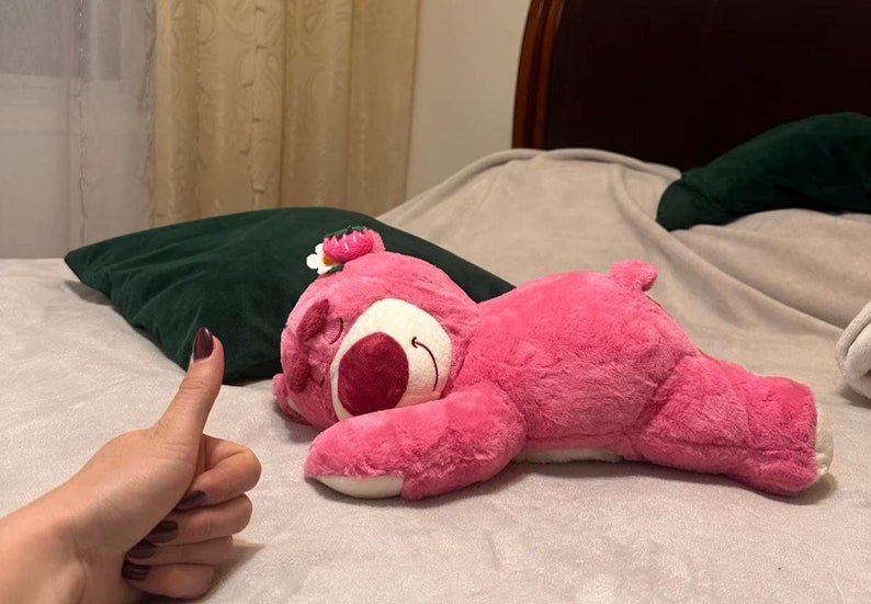May include: A pink plush bear toy with a white belly and a flower on its head is lying on a bed. The bear has a smiling face and is in a relaxed position. A dark green cushion is behind the bear.