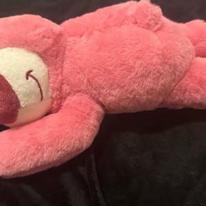 May include: A pink plush bear toy with a white face and a dark red nose. The bear has a small strawberry and flower accessory on its head. The bear is lying down on a dark surface.