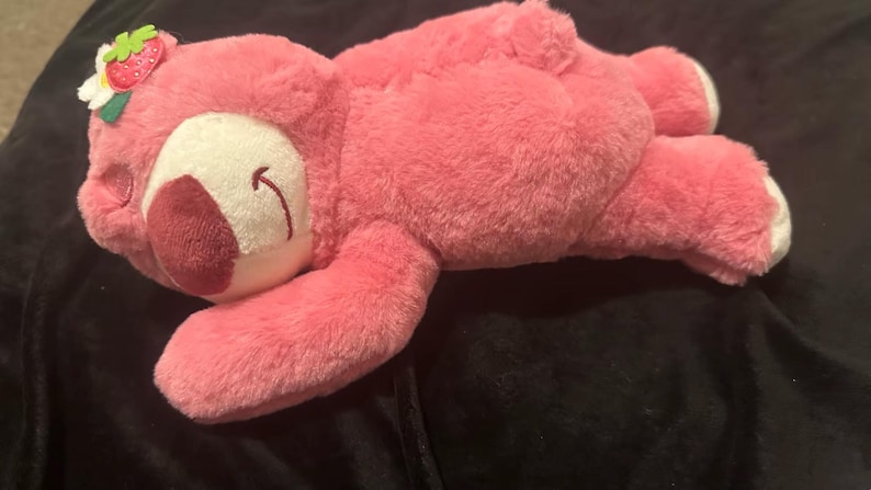 May include: A pink plush bear toy with a white face and a dark red nose. The bear has a small strawberry and flower accessory on its head. The bear is lying down on a dark surface.