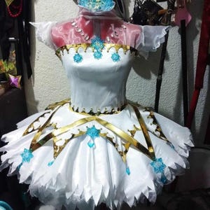 Könnte beinhalten: Weißes Cosplay-Kleid mit goldenen Verzierungen und blauen Akzenten. Das Kleid hat ein Oberteil, einen mehrlagigen Rock und ein passendes Kopfstück. Das Design umfasst goldfarbene kronenförmige Verzierungen und blaue kristallartige Details.
