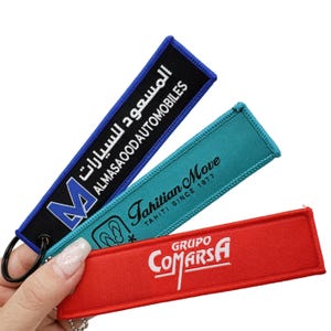 May include: Three embroidered keychains in blue, teal, and red. The blue keychain has Arabic text and "ALMASAOOD AUTOMOBILES". The teal keychain reads "Tahitian Move TAHITI SINCE 1977". The red keychain says "GRUPO COMARSA".