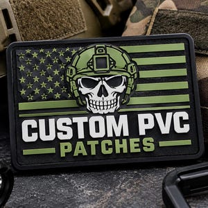 Puede incluir: Un parche rectangular de PVC con un diseño estilizado de la bandera estadounidense en negro y verde oliva. Centrado en la bandera hay una calavera blanca con un casco táctico verde oliva. El texto CUSTOM PVC PATCHES aparece en color blanco.