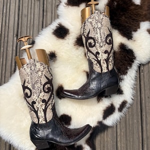 May include: A pair of cowboy boots with a snakeskin pattern on the upper shaft and decorative stitching. The boots are black with a pointed toe and a low heel. They are displayed on a white and brown patterned rug.